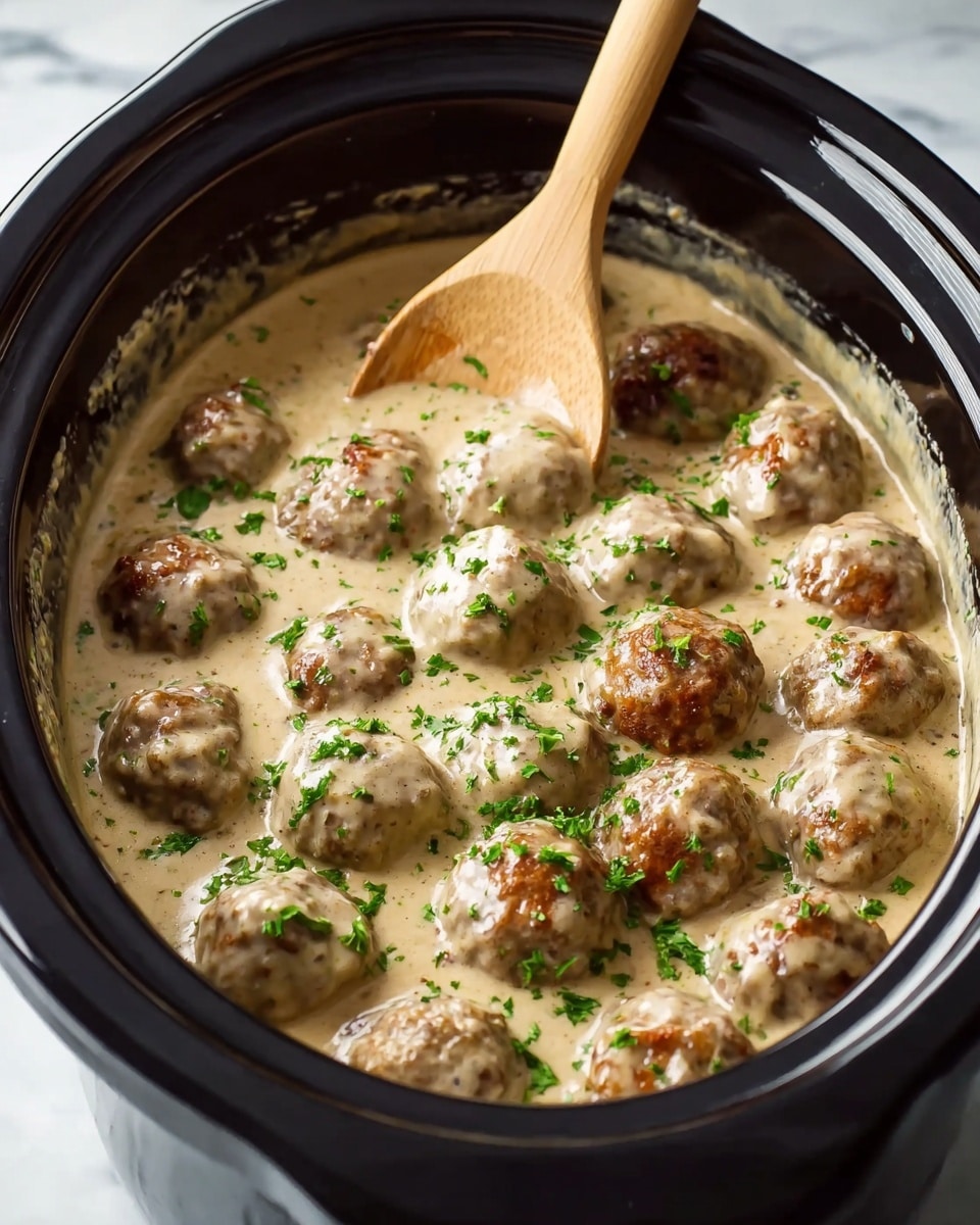 The image shows a dark grey bowl filled with wide, curly egg noodles at the bottom layer, which are light yellow with a smooth texture. On top of the noodles, there are around fifteen round meatballs covered in thick, creamy light brown sauce, some with browned, slightly crispy spots. The meatballs are sprinkled with finely chopped green herbs. A silver fork is placed on the right side of the bowl. The scene is set on a white marbled surface. photo taken with an iphone --ar 4:5 --v 7
