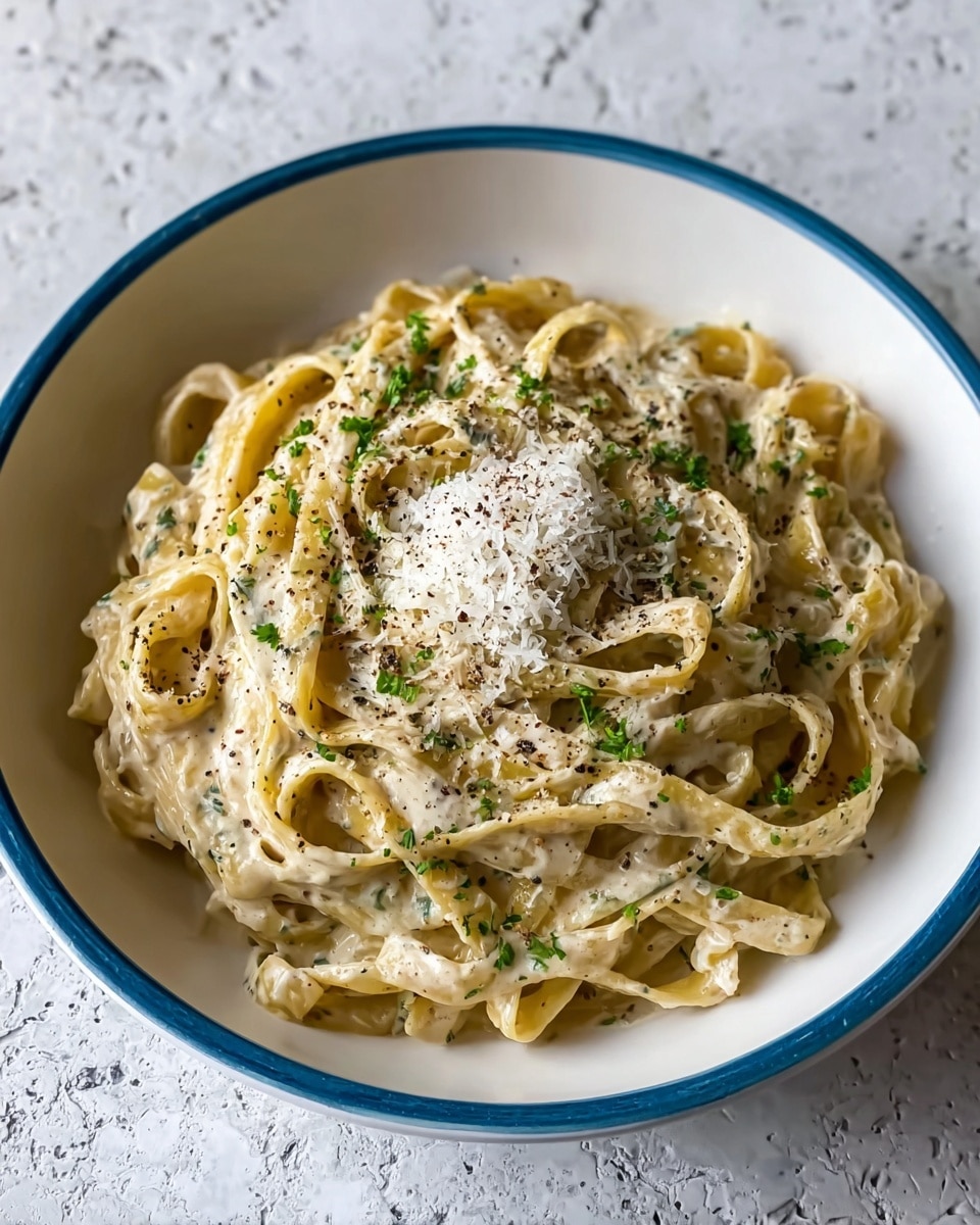 The image shows a white bowl filled with creamy fettuccine pasta. The pasta strands are coated in a thick, smooth light yellow sauce speckled with small green herb pieces. The top of the pasta has a small pile of finely grated white cheese and a light sprinkle of cracked black pepper. The bowl sits on a white marbled surface, and in the background, there is a dark blue pot slightly out of focus. The lighting highlights the glossy texture of the sauce and the strands of pasta. Photo taken with an iphone --ar 4:5 --v 7