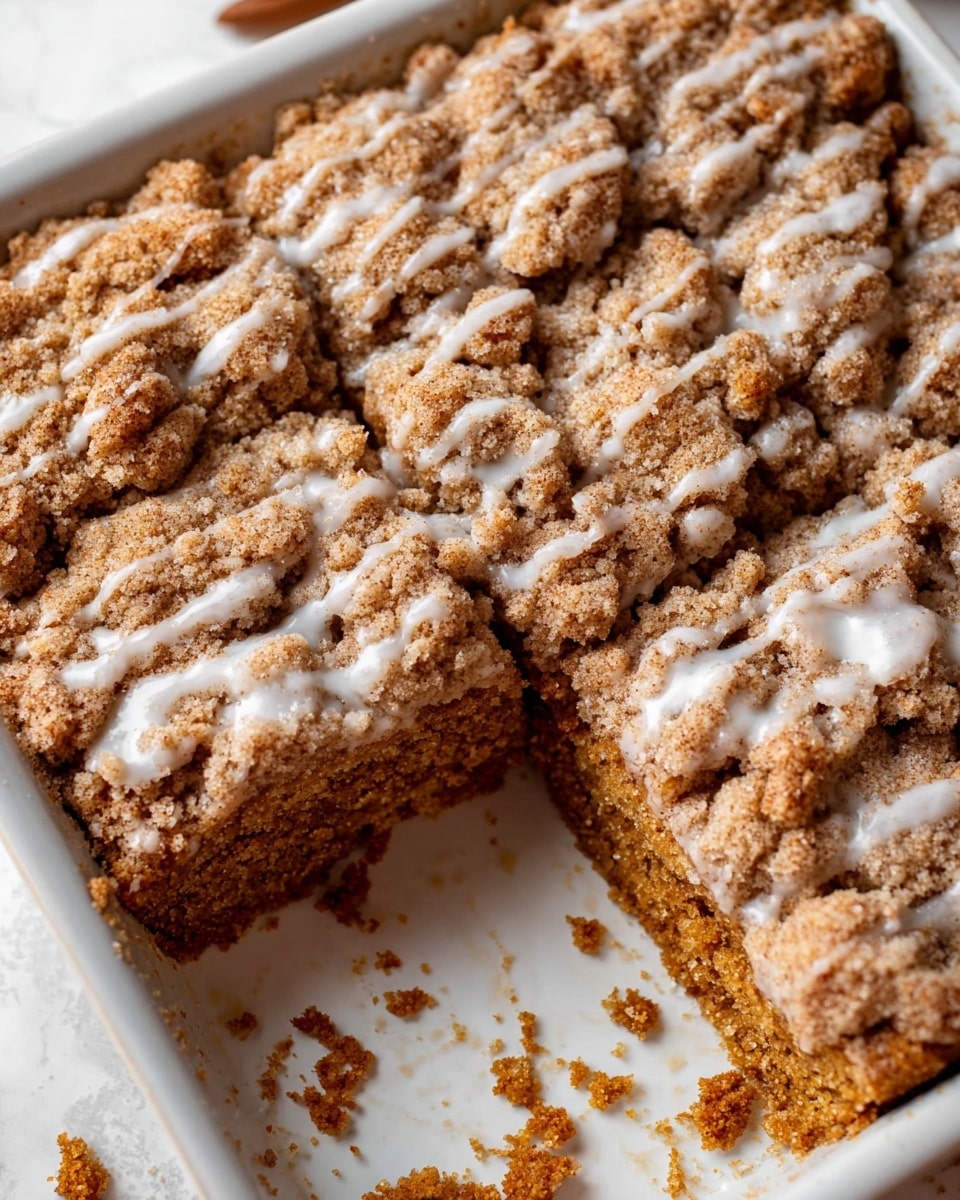 The dish is a square-shaped crumb cake in a white baking dish with a crumbly top layer that is golden brown with rough, chunky crumbs, drizzled with a light white glaze. The middle layer is moist and dense with a warm brown color, showing a soft, fine texture. The crumb topping covers the entire surface, with one square piece removed, revealing the thick, spongy cake underneath. Small crumbs are scattered around the empty space on the white marbled surface underneath the dish. photo taken with an iphone --ar 4:5 --v 7