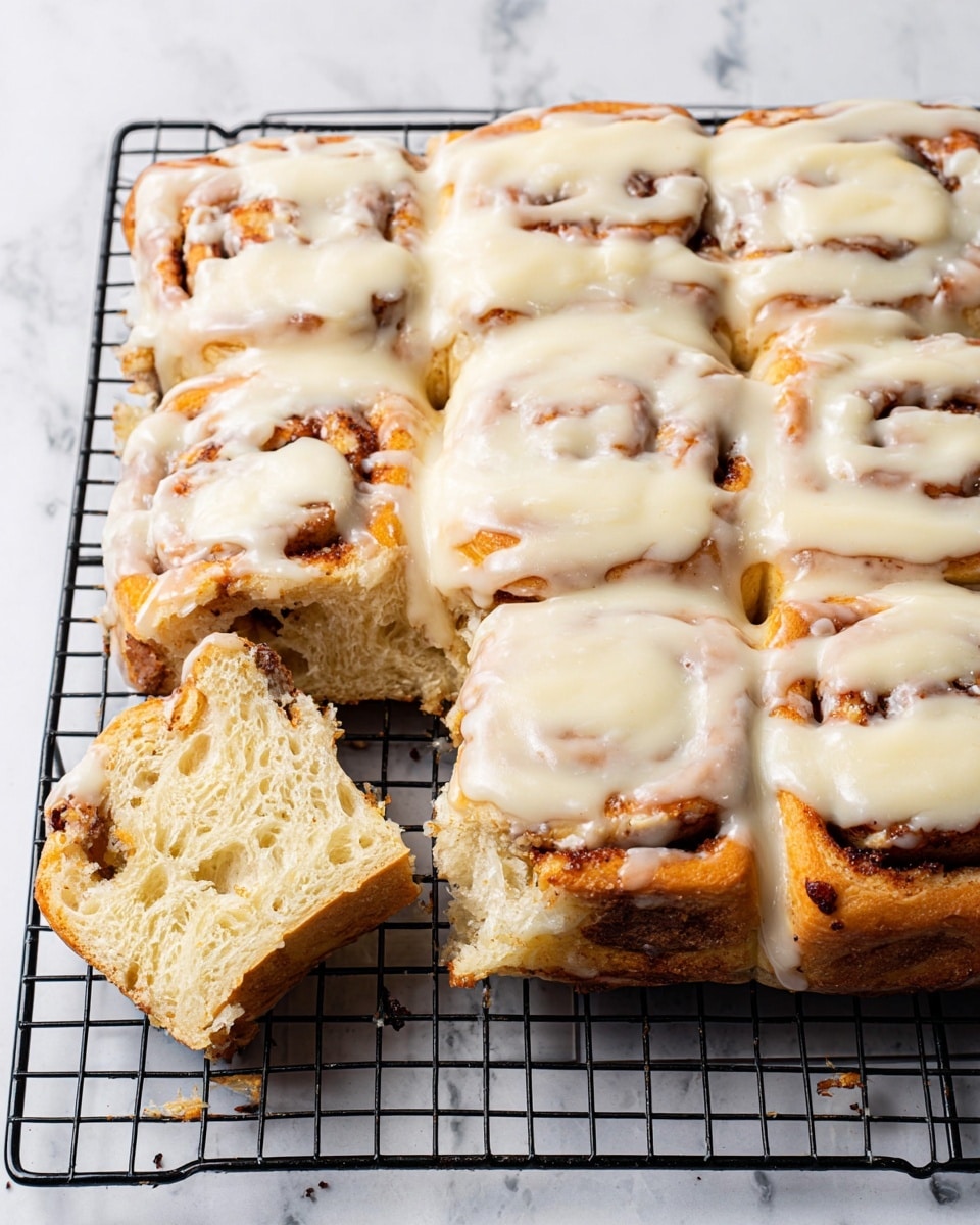 The image shows a rectangle of cinnamon rolls covered with creamy white icing on top, resting on a black wire cooling rack over a white marbled surface. The cinnamon rolls are visible underneath the thick, smooth icing with uneven patches showing the brown cinnamon swirls beneath. The icing looks soft and slightly dripping over the edges of the rolls. The rolls are arranged close together, filling the whole rectangle with a golden-brown color visible through the icing. Photo taken with an iphone --ar 4:5 --v 7