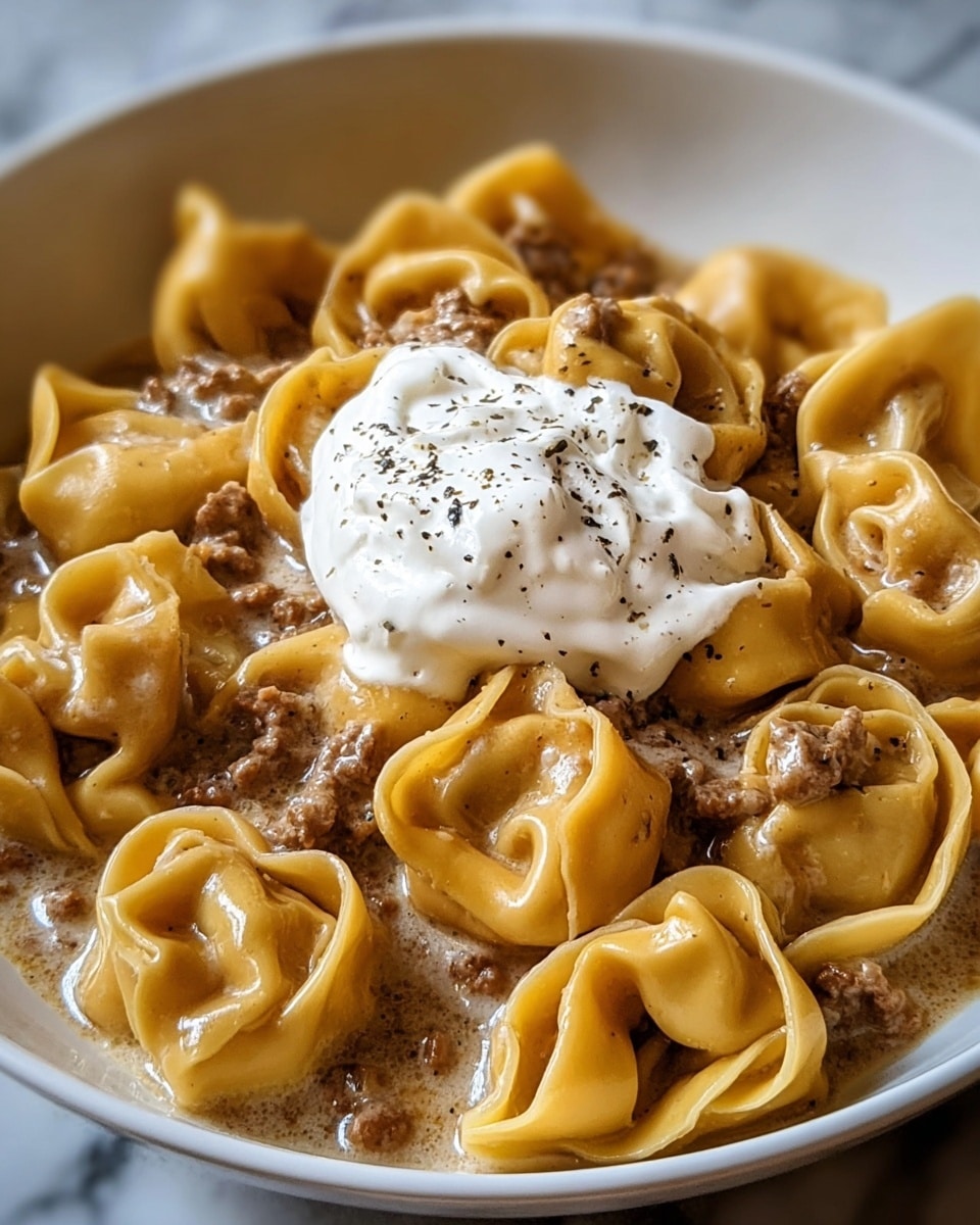 A white bowl filled with a creamy pasta dish that has many tortellini pasta pieces, each stuffed with brown meat and coated in a light brown creamy sauce. On top, there is a dollop of white sour cream sprinkled with small black pepper bits. The tortellini pasta looks soft and smooth with a yellowish color. The bowl is placed on a white marbled surface. photo taken with an iphone --ar 4:5 --v 7