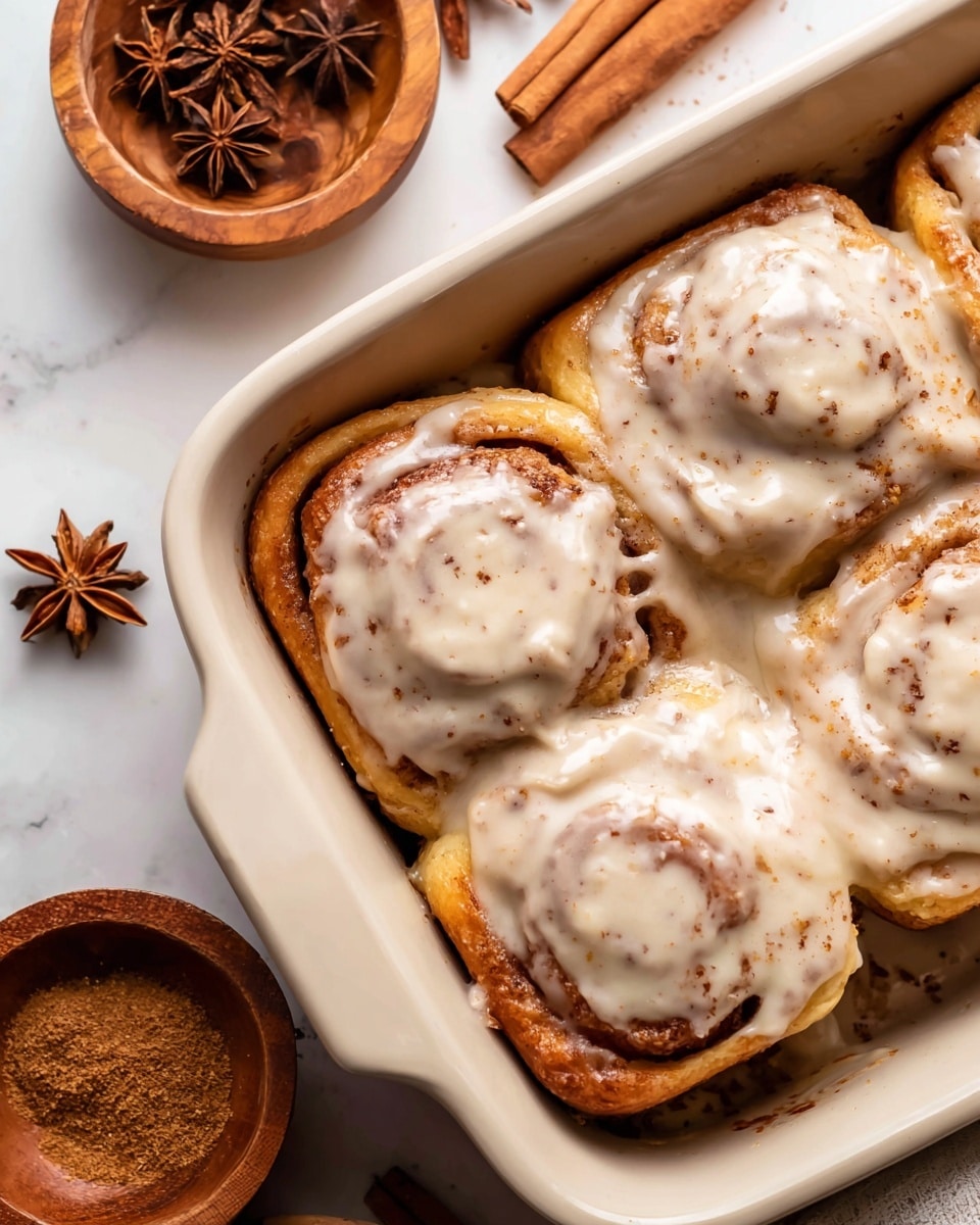 The image shows a close-up view of several cinnamon rolls covered in a thick, creamy light brown frosting with specks of cinnamon visible throughout. Each roll has a distinct spiral pattern with the frosting filling the spaces between the layers, giving a swirled texture. There are a few whole star anise pods placed on top, adding a dark brown star-shaped contrast against the lighter frosting. The rolls are set closely together, filling the frame with their soft and smooth texture. The background is a white marbled surface. Photo taken with an iphone --ar 4:5 --v 7