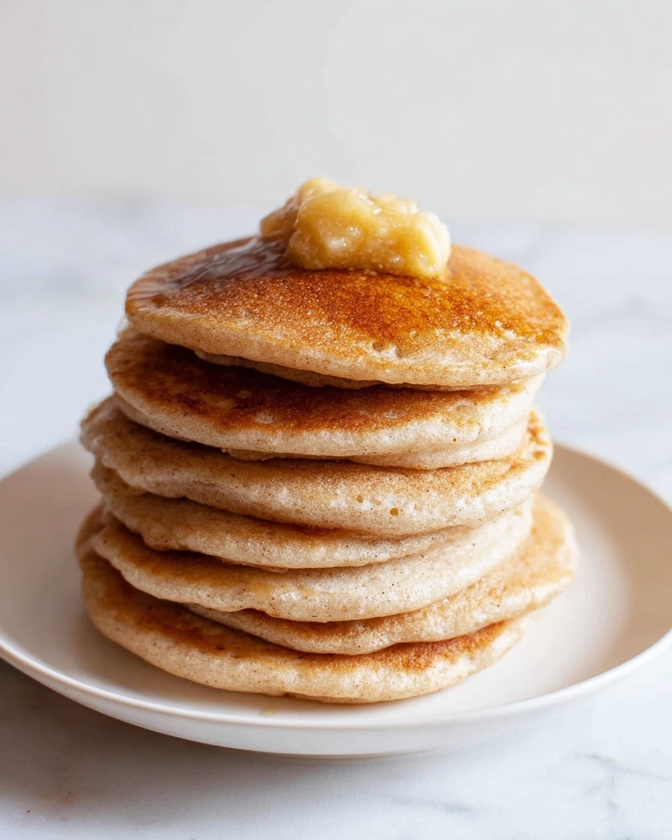 A white plate holds seven round pancakes, each with a golden-brown top surface and a light beige underside, arranged in a loose circle with some slightly overlapping. The pancakes have a soft, fluffy texture with small air holes visible on their sides. In the center of the plate sits a small dollop of mashed yellow banana, adding a slightly chunky texture and a pop of color. The plate rests on a white marbled surface. photo taken with an iphone --ar 4:5 --v 7