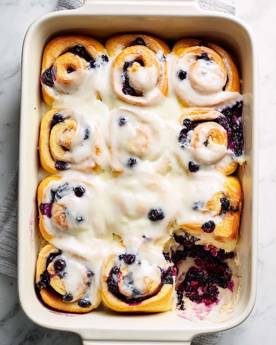 Large white rectangular baking dish filled with a full batch of freshly baked blueberry cinnamon rolls generously covered in creamy white icing, soft golden-brown spiral dough swirled with vibrant whole blueberries peeking through, the entire dish photographed from a 3/4 angle on a white marble countertop with natural lighting, styled for a food magazine hero shot, photo taken with an iphone --ar 4:5 --v 7