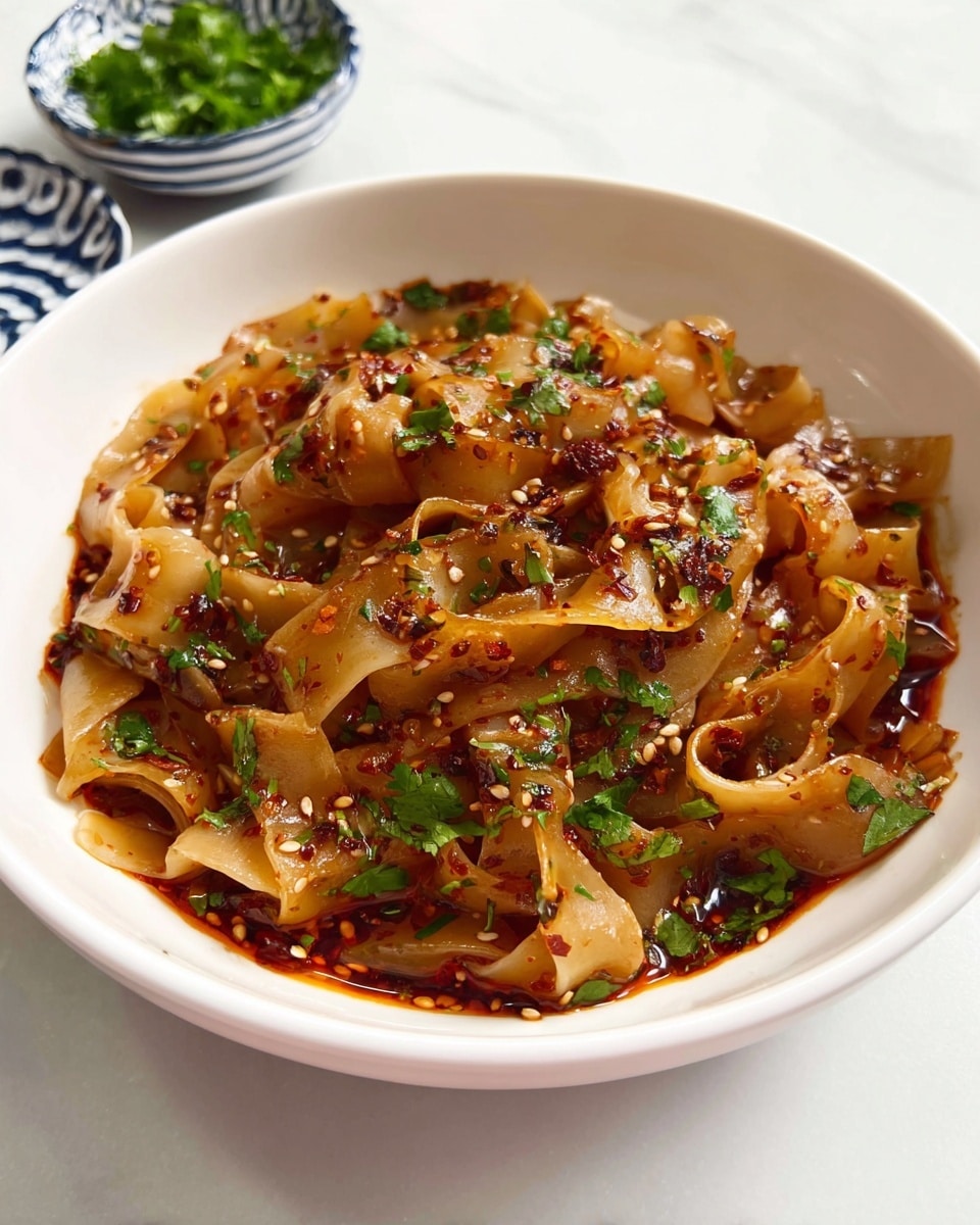 Large white serving bowl filled with the entire portion of wide, glossy hand-pulled noodles tossed in a vibrant, spicy chili oil sauce, garnished with scattered toasted sesame seeds, fresh chopped cilantro, and green onions, aromatic sauce pooling slightly at the bottom, presented with small white bowls of fresh herbs on the side, all photographed from a professional 3/4 angle on white marble surface with natural lighting, resembling a hero shot from a food magazine, photo taken with an iphone --ar 4:5 --v 7