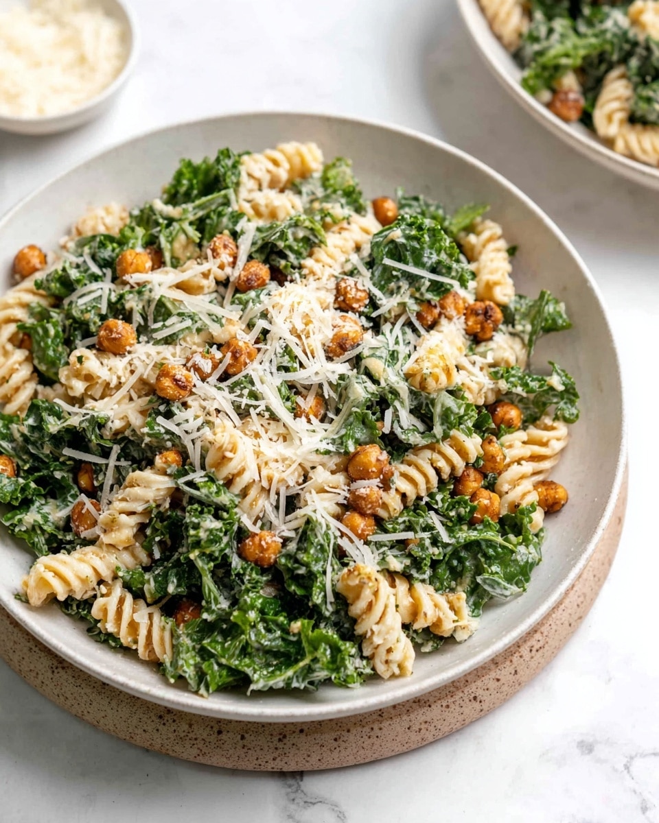 Large white bowl filled with creamy kale pasta salad featuring spiral rotini pasta mixed evenly with fresh green kale leaves and crunchy roasted chickpeas, generously topped with shredded parmesan cheese, presented on a white marble countertop with natural lighting, professional 3/4 angle food magazine hero shot showing the entire dish in frame, photo taken with an iphone --ar 4:5 --v 7