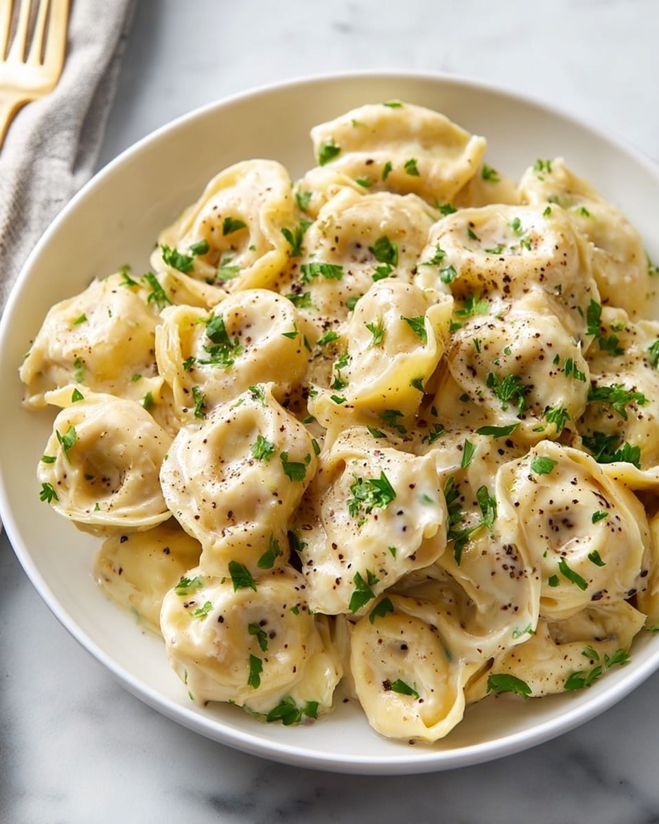 Large white serving bowl filled with a generous portion of creamy cheese tortellini pasta, evenly coated in a rich, velvety Alfredo sauce speckled with freshly cracked black pepper and finely chopped parsley, entire dish presented from a 3/4 angle on a pristine white marble countertop, styled with natural lighting for a fresh and appetizing look, professional food magazine hero shot photo taken with an iphone --ar 4:5 --v 7