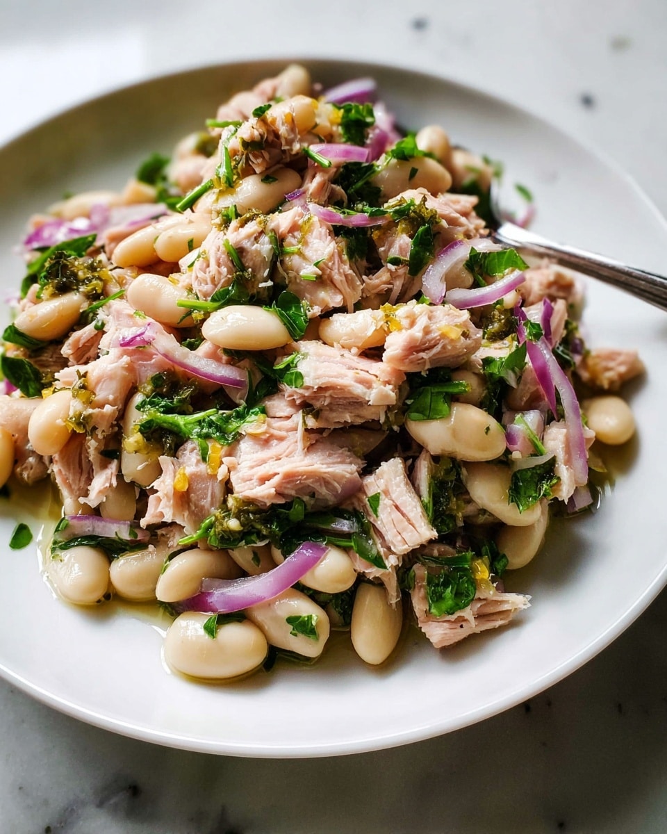 Large white serving bowl filled with a vibrant tuna and white bean salad, featuring tender chunks of tuna mixed with plump white beans, finely chopped red onions, and fresh green herbs like parsley, all lightly dressed with olive oil and lemon zest, whole dish displayed beautifully on a white marble countertop, natural lighting highlighting the fresh textures and colors, professional food styling photo taken with an iphone --ar 4:5 --v 7