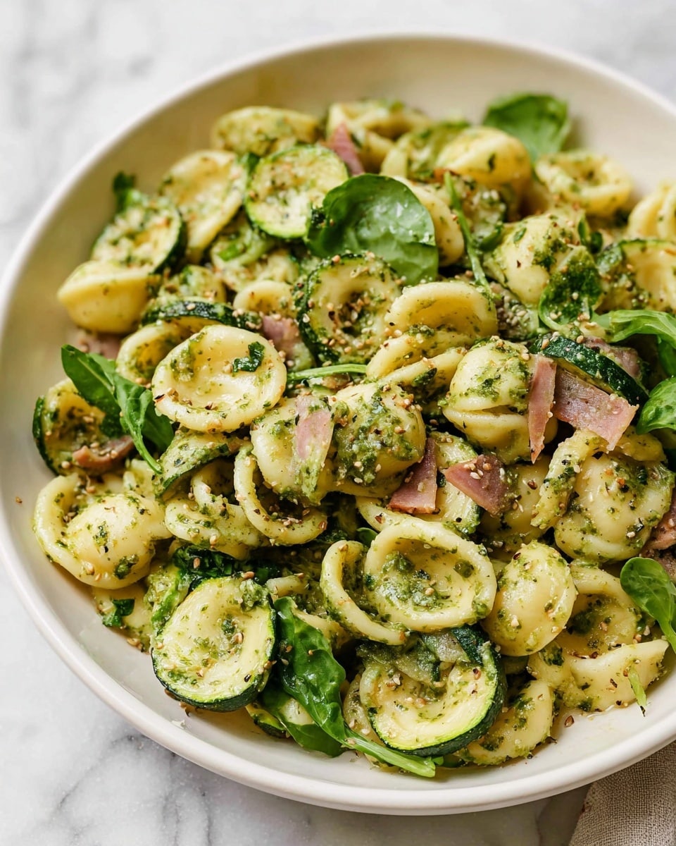 Large white bowl filled with a generous serving of orecchiette pasta tossed in a vibrant green pesto sauce, mixed with grilled zucchini slices, fresh spinach leaves, and diced red onions, all evenly coated and glistening with herbs, presented from a 3/4 angle on a white marble countertop, natural lighting highlighting the fresh ingredients, professional food styling photo taken with an iphone --ar 4:5 --v 7
