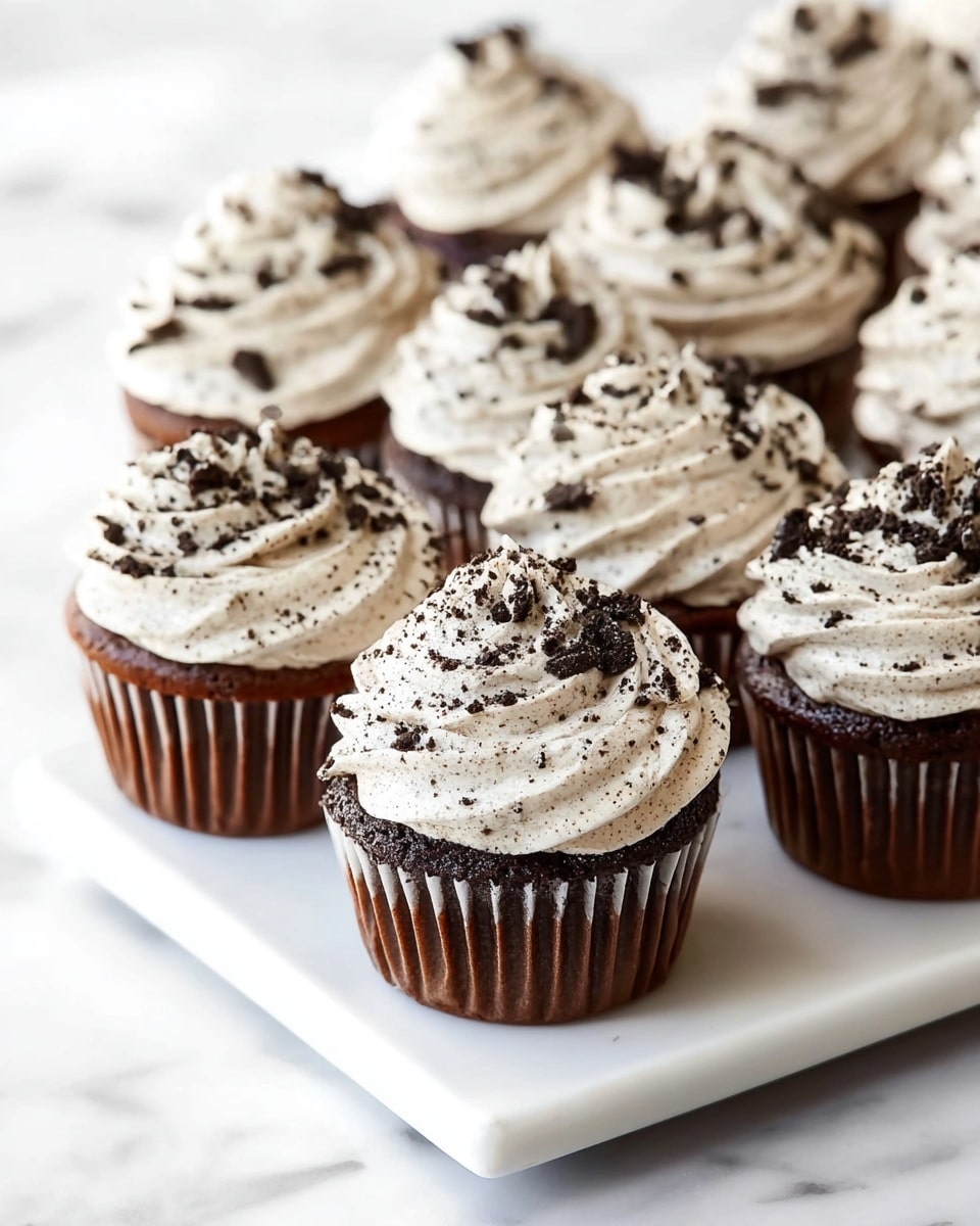 A white rectangular platter displaying a complete batch of rich chocolate cupcakes, each topped generously with a thick layer of creamy cookies and cream frosting speckled with crushed chocolate cookie bits, arranged closely together to showcase their uniform decoration and texture, set on a white marble countertop with natural lighting highlighting the soft frosting waves and chocolate cake bases, professional 3/4 angle food magazine hero shot, photo taken with an iphone --ar 4:5 --v 7